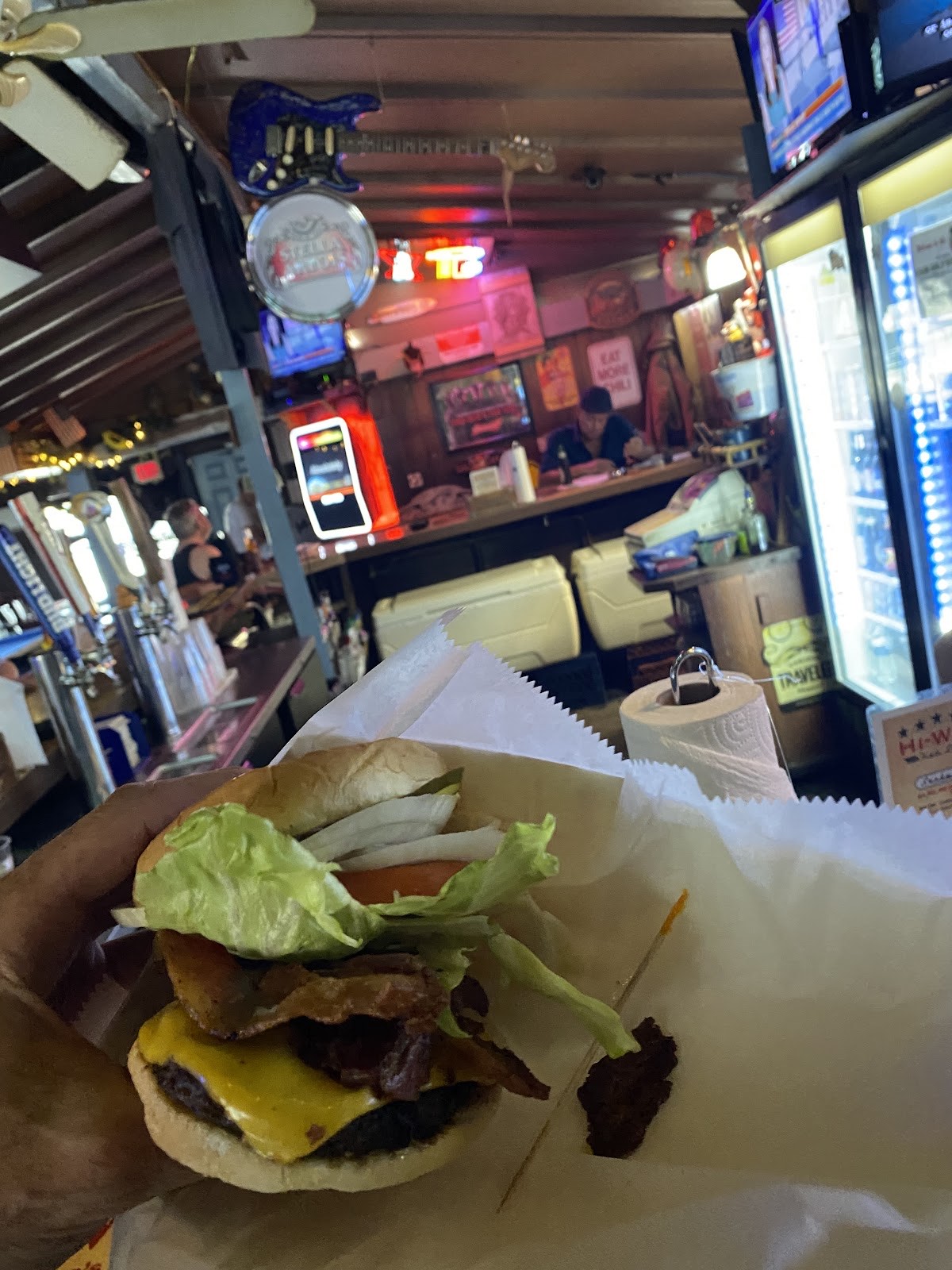A loaded bacon cheeseburger held up inside Hi Way Bar with neon beer signs glowing behind the bar