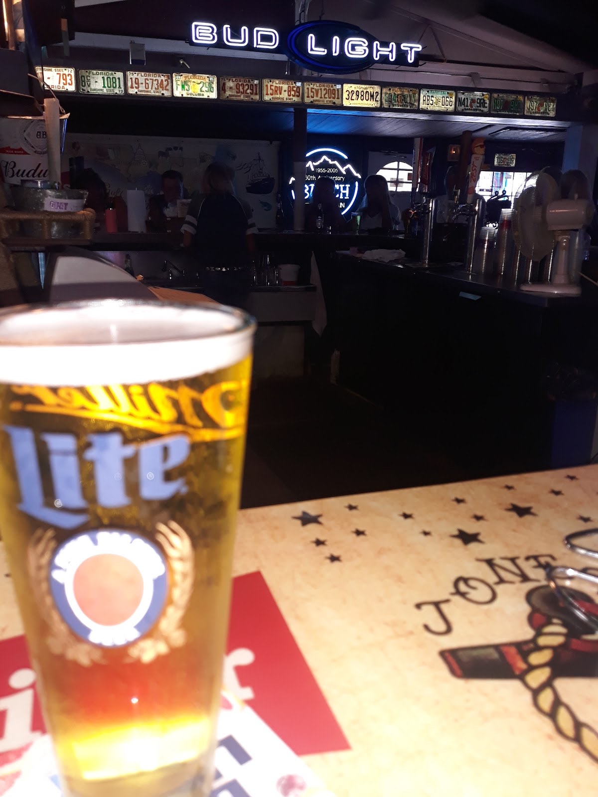 A frosty Miller Lite pint glass on the bar with a glowing Bud Light neon sign and license plates behind