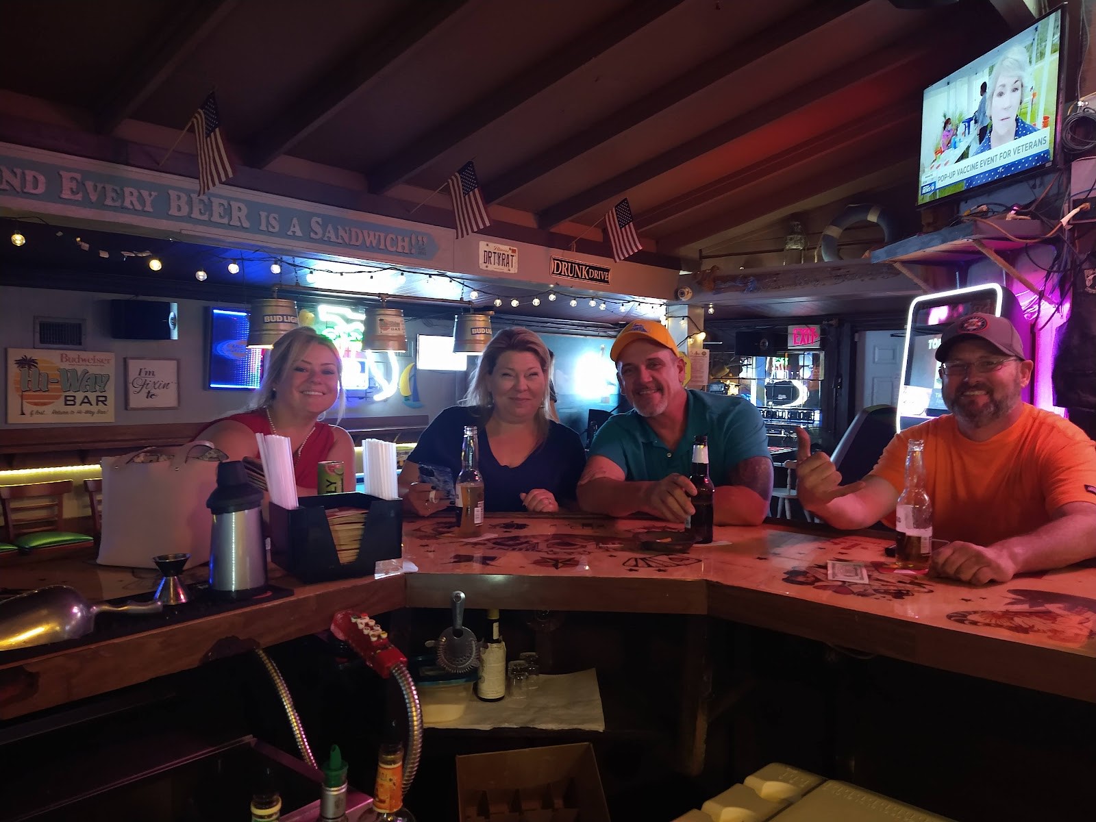 Friendly regulars raising bottles at the bar inside Hi Way Bar under a sign reading Every Beer is a Sandwich