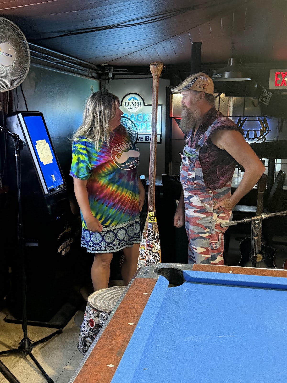 Two patrons chatting by the pool table and arcade game inside the dimly lit dive bar